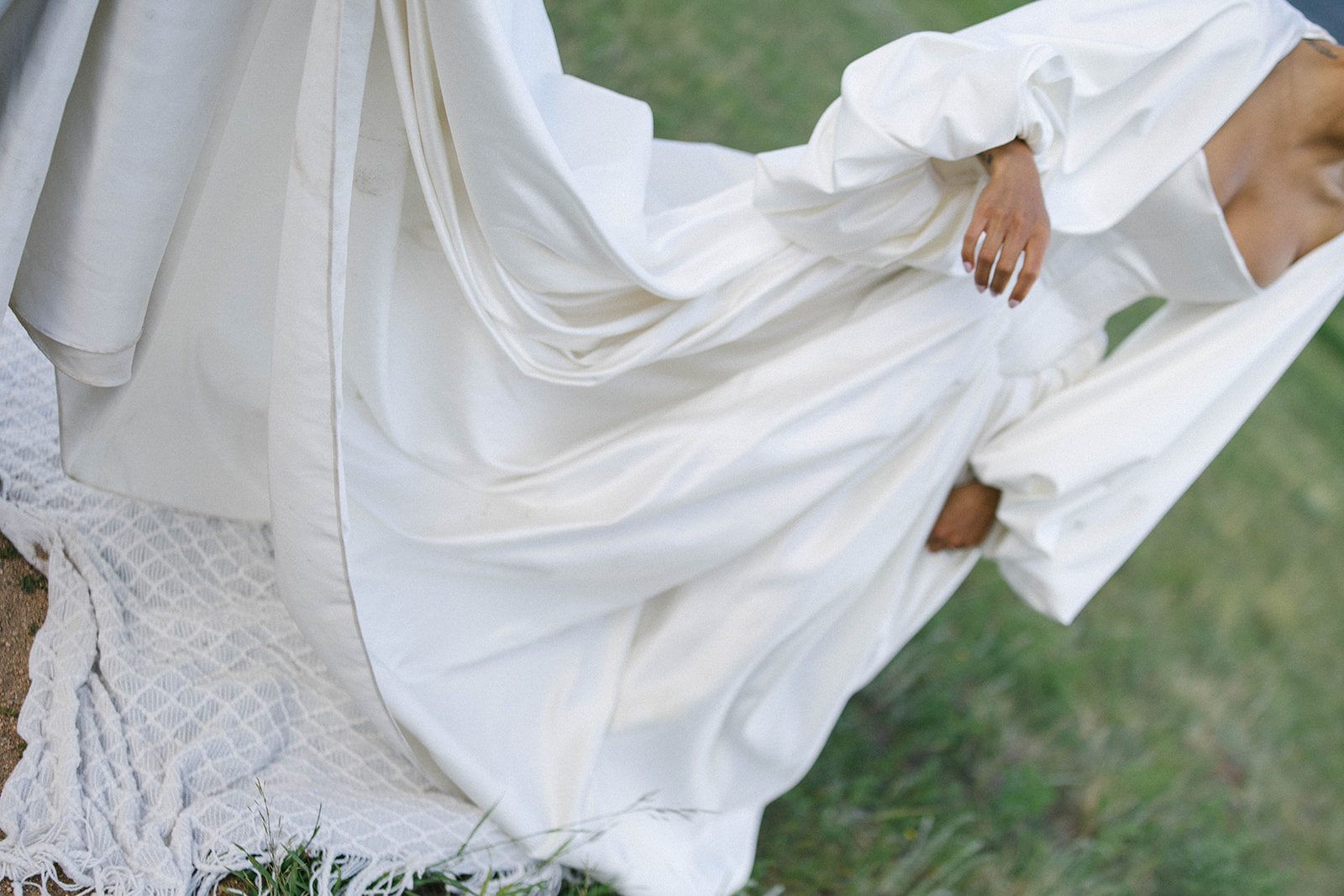 Detail shot of a bride’s gown, the folds of voluminous white fabric spilling across a patterned blanket during their outdoor celebration.