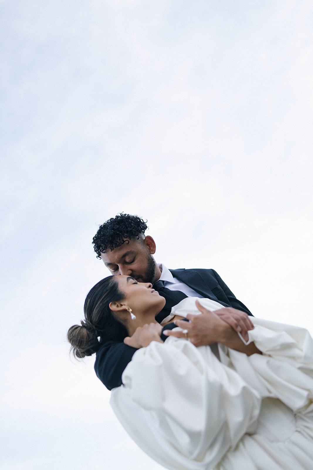 Close-up of a groom kissing his bride’s forehead as she leans into him, her flowing white gown standing out against the soft blue sky. Captured by a Colorado wedding photographer.
