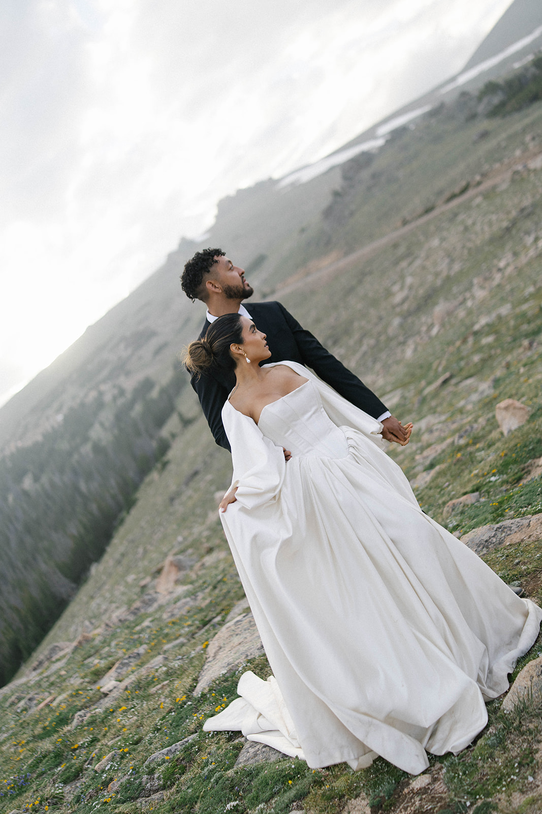 A bride in a flowing white gown holds her partner’s hand as they stand on a rocky mountainside, looking out over the sweeping Colorado landscape. Captured by a Colorado wedding photographer, the moment feels cinematic and intimate.
