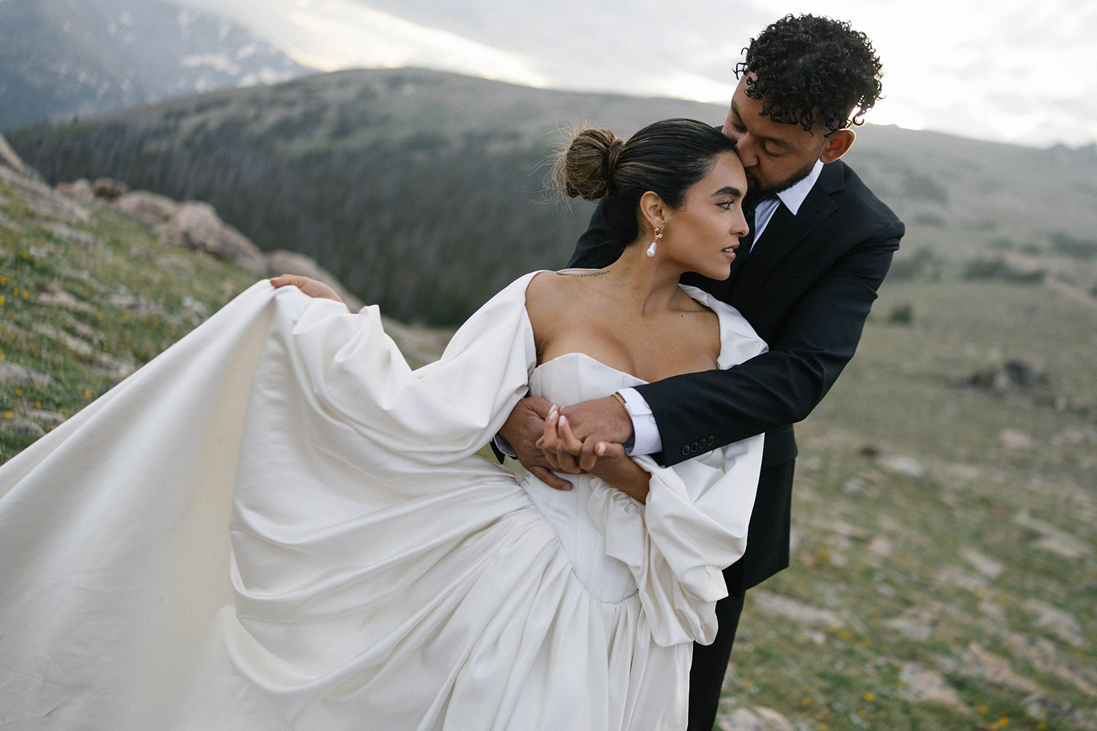 Romantic mountain embrace as the groom holds his bride close, her elegant gown billowing as he kisses her forehead with alpine views behind them. Captured by a Colorado wedding photographer.