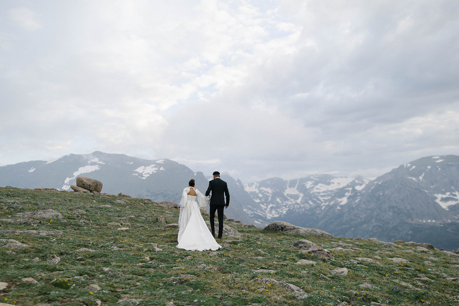 A couple in wedding attire walks hand in hand across a high alpine meadow with snow-capped peaks in the distance, captured by a Colorado wedding photographer.