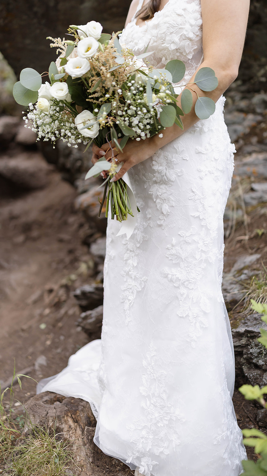 Bride in a lace wedding gown holding a lush bouquet of white roses, eucalyptus, and baby’s breath in a mountain setting.