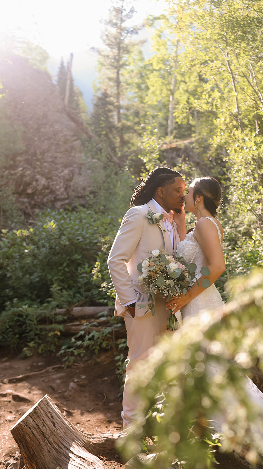 Couple sharing a kiss in the forest with golden sunlight streaming through the trees, the bride holding a bouquet of greenery and white florals