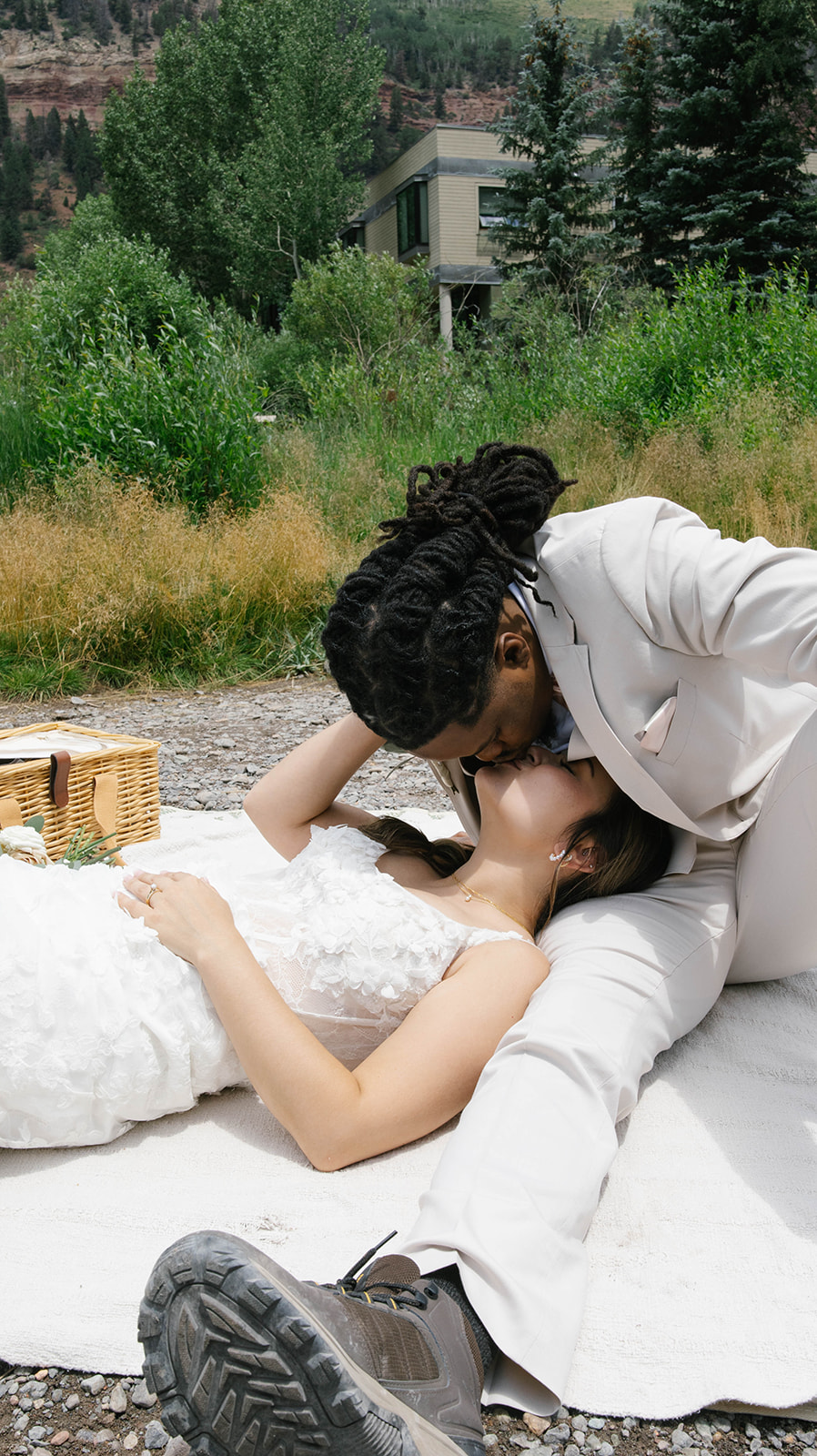 A couple reclines on a picnic blanket, the groom leaning down for a kiss while the bride lays back smiling, surrounded by tall summer grasses.