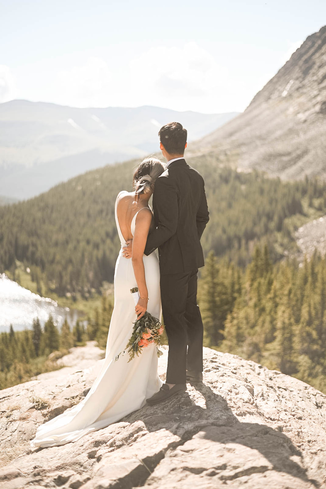 Bride and groom standing on a cliffside, arms wrapped around each other, looking out over the Colorado mountains.
