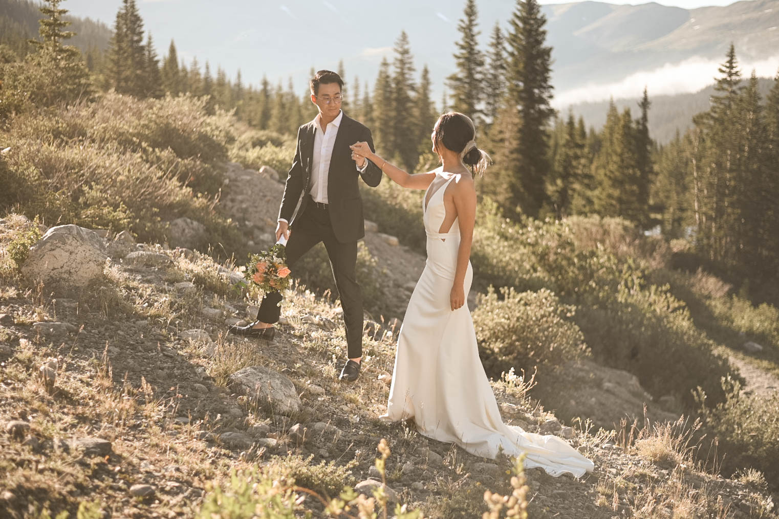 A groom in a dark suit helps his bride in a sleek white gown walk across rocky terrain at golden hour, surrounded by pine trees.