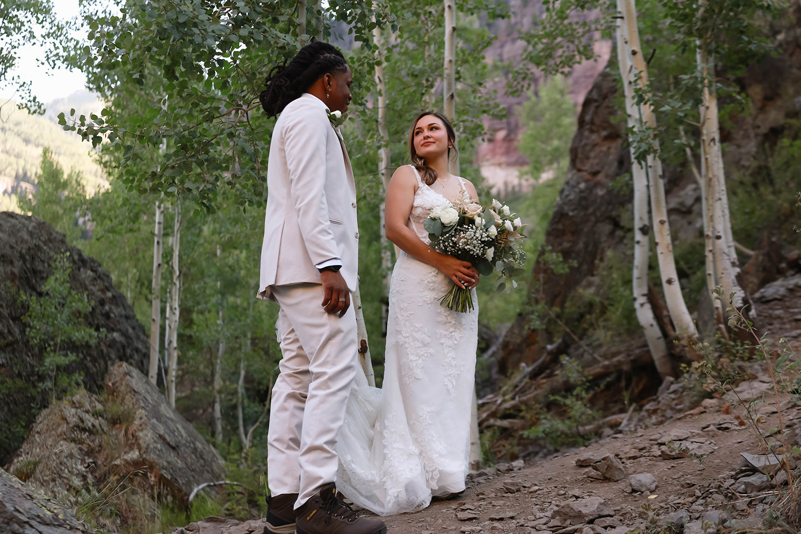 The couple stands together on a rocky mountain path surrounded by aspen trees, sharing a quiet, romantic moment in the forest.