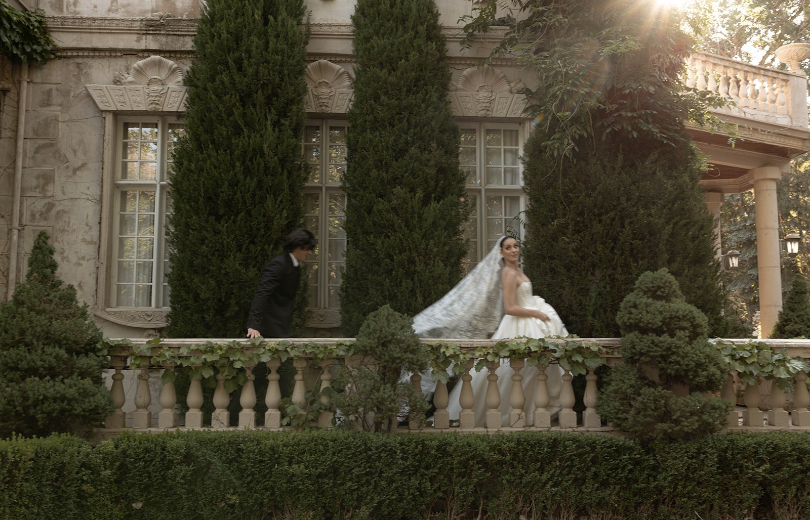 The bride glances back as her groom follows along an ivy-covered terrace framed by tall cypress trees at La Caille, Utah.
