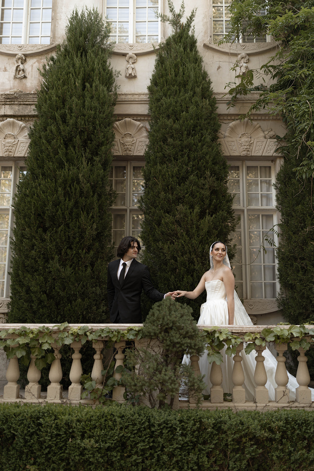 The couple stands hand in hand between lush greenery and ornate architecture, capturing a timeless European-inspired moment.