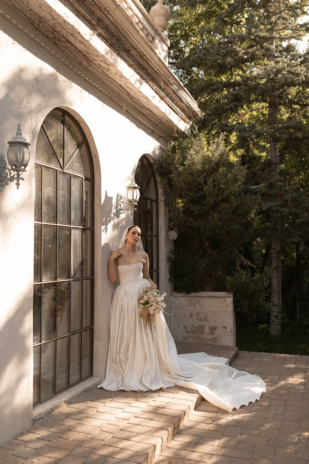 The bride leans into the warm light, lace veil draped around her, bouquet in hand, creating a serene and romantic portrait.
