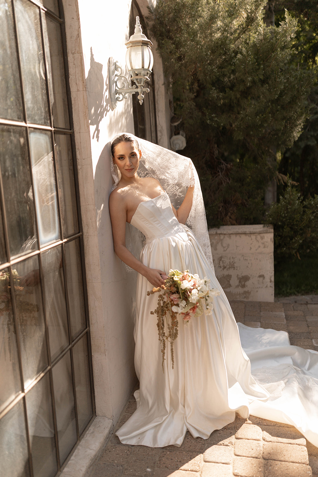 The bride stands elegantly by the sunlit arched windows of La Caille, Utah, holding a cascading pastel bouquet