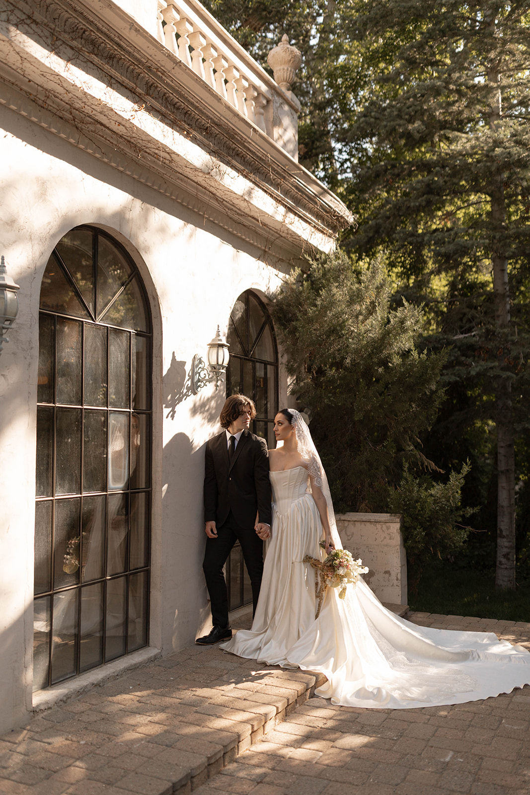 The couple stands close beside the vintage stone walls of La Caille, Utah, the bride’s long train trailing down the steps.
