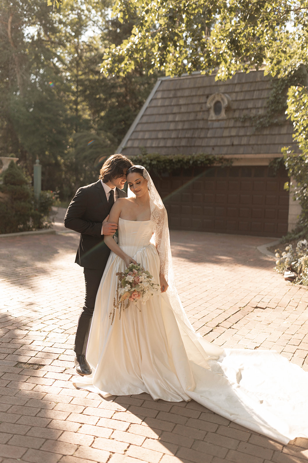 The bride and groom share a quiet embrace in golden sunlight, surrounded by garden greenery and cobblestone texture.
