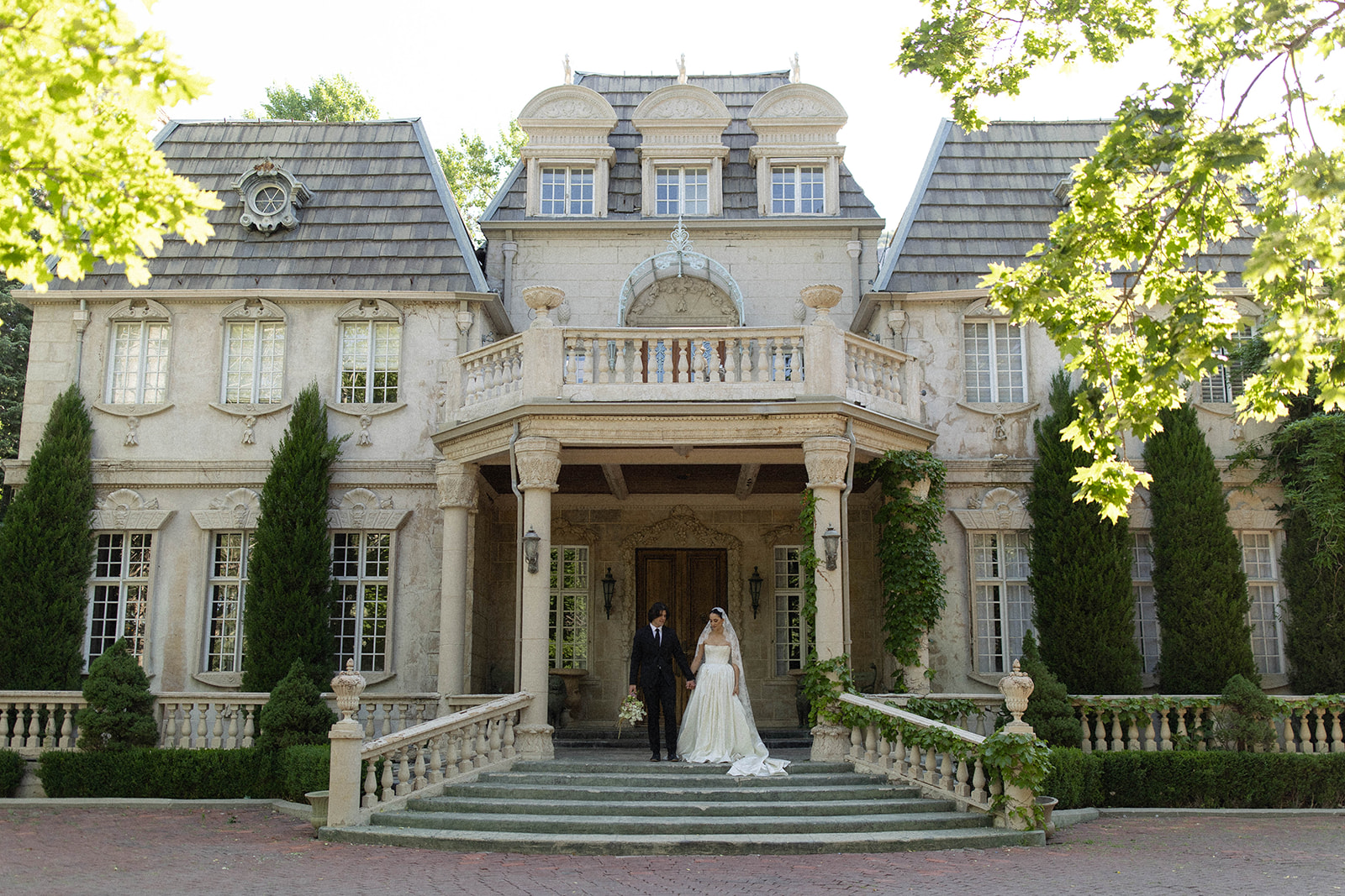 The bride and groom walk hand in hand down the cobblestone driveway in front of the grand French-style mansion at La Caille, Utah, framed by soft morning light and lush greenery.