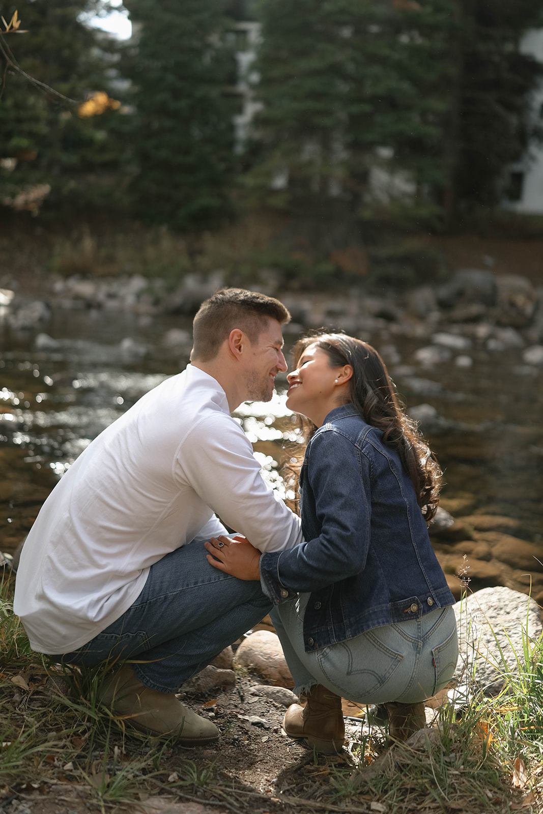 A couple crouches by a rocky riverbank, leaning in for a kiss as sunlight hits the water in vail village in Colorado.