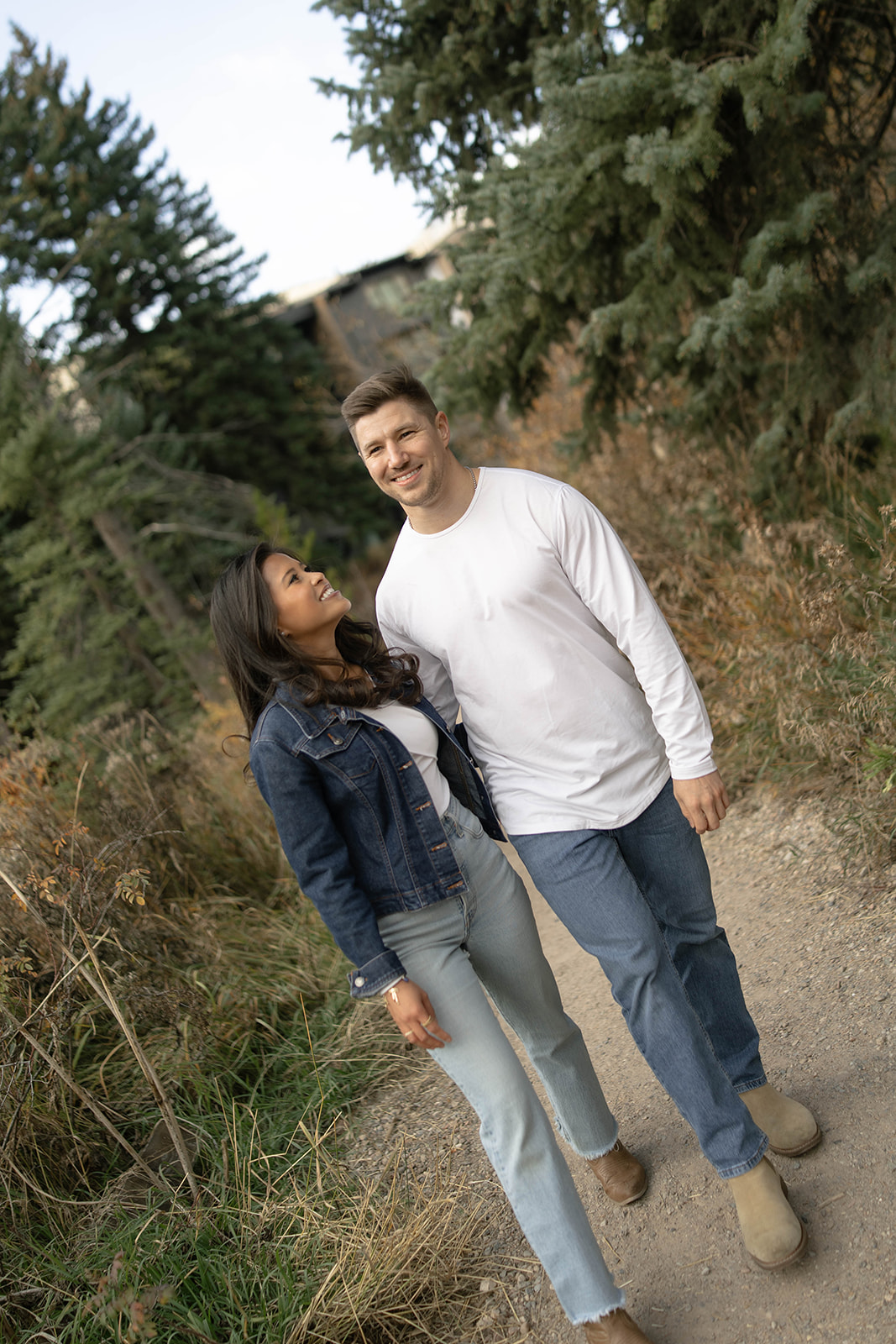 A couple strolls down a forested trail, smiling warmly at each other with tall evergreens in the background.