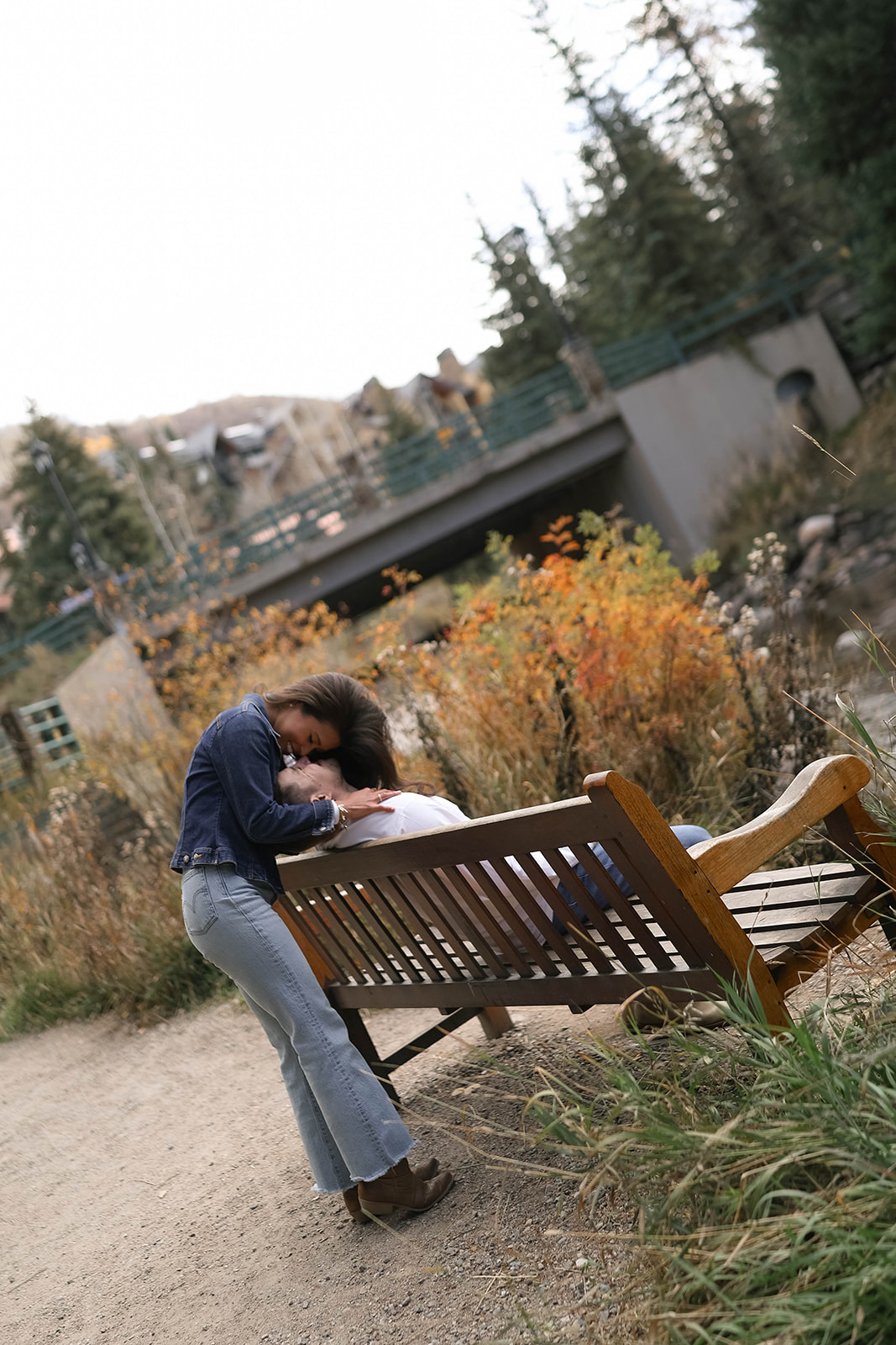 A woman leans over a wooden bench to kiss her partner, with fall foliage and a small bridge near vail village in Colorado.