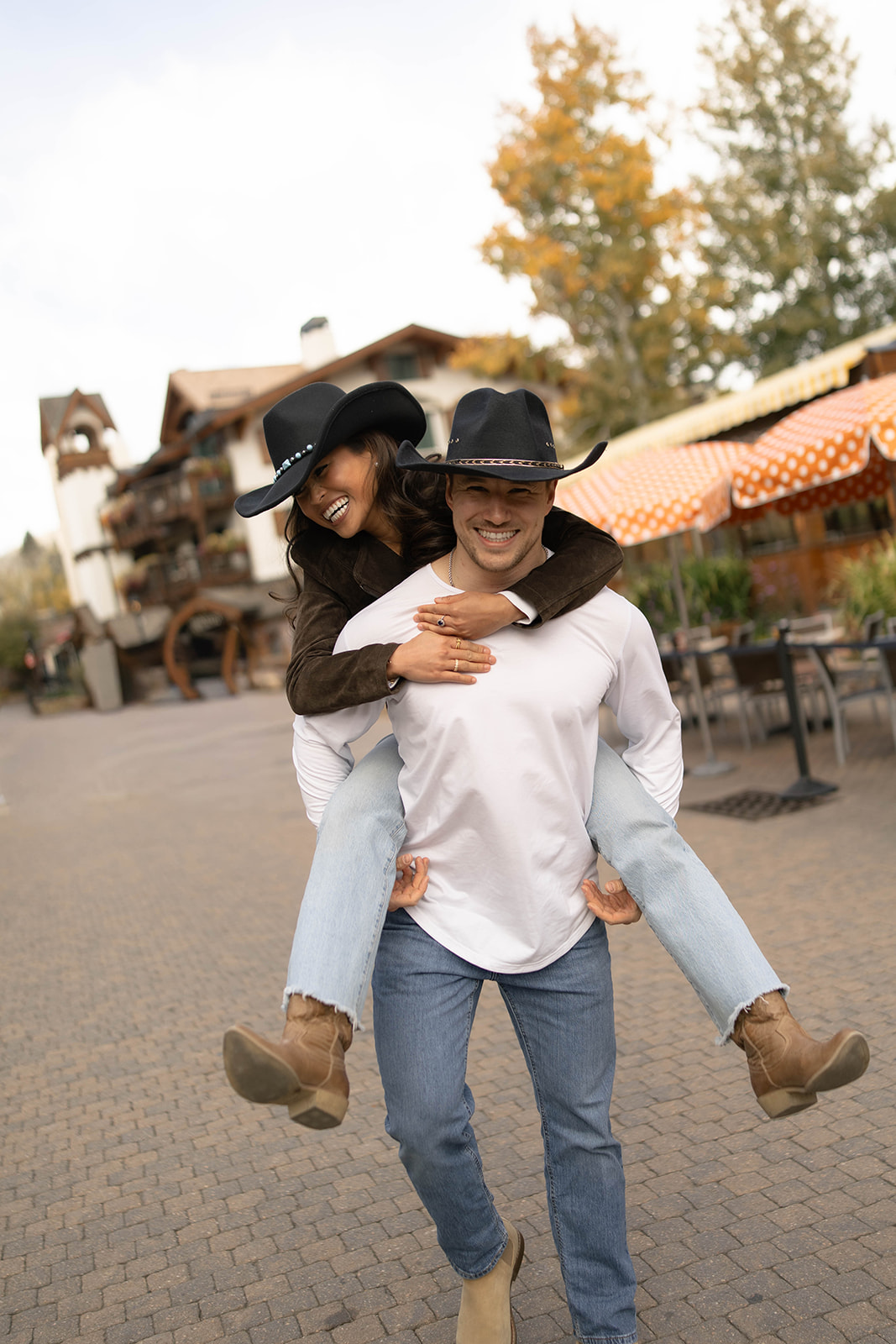 Couple laughing as she rides on his back while exploring Vail Village in Colorado in their matching cowboy hats.