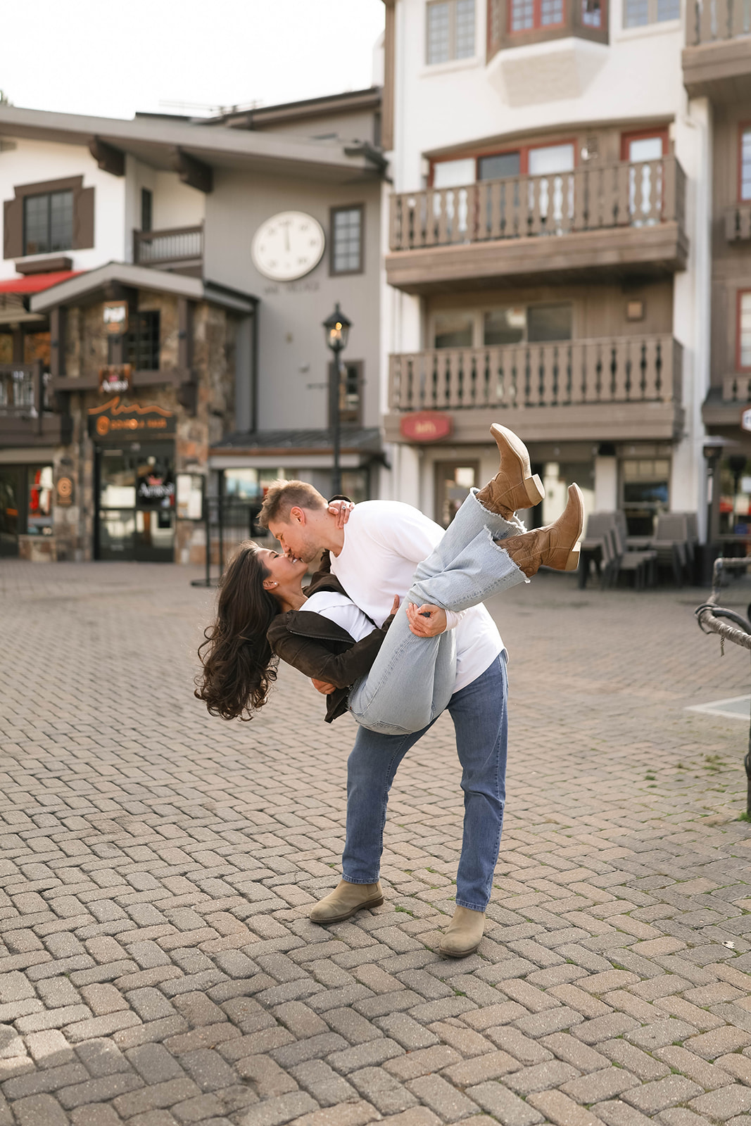 A man lifting his partner in a playful dip as they kiss in a cobblestone plaza in Vail Village in Colorado, with rustic shops and balconies behind them.
