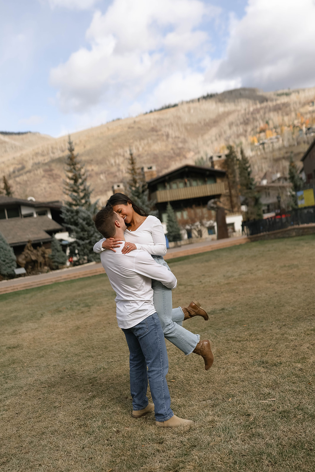A man lifts his partner playfully in an open grassy field with mountains rising behind them.