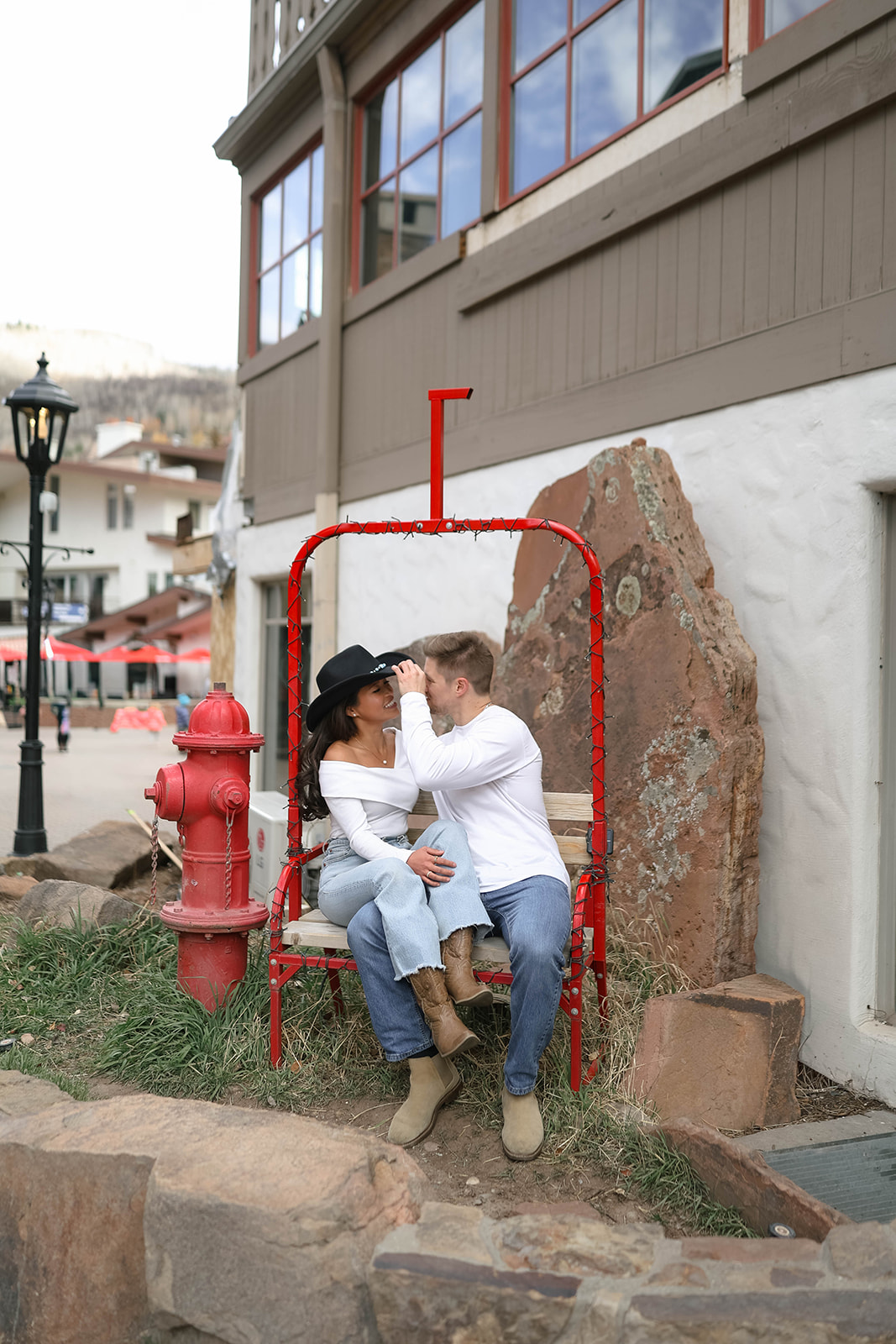 Couple sitting on a red lift-style bench, playfully adjusting her hat while enjoying a cozy moment in Vail Village in Colorado.