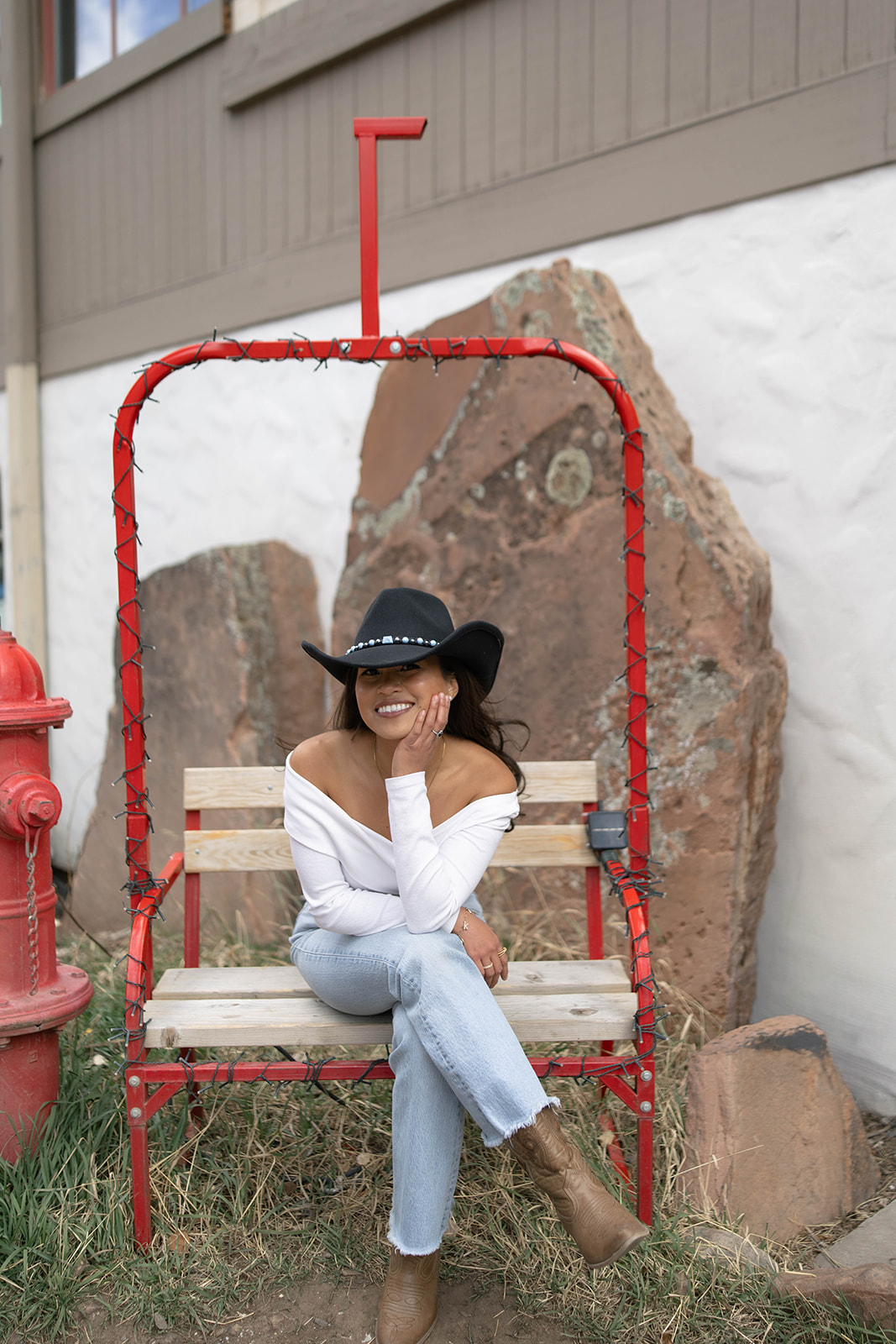 A woman in a white off-the-shoulder top and cowboy hat sitting on a red ski-lift style bench, smiling with her hand resting on her cheek.