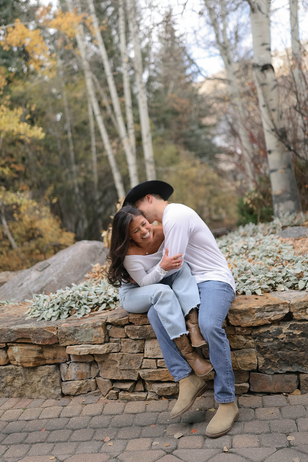 A couple laughing together as they cuddle on a stone wall surrounded by autumn foliage and tall aspen trees.