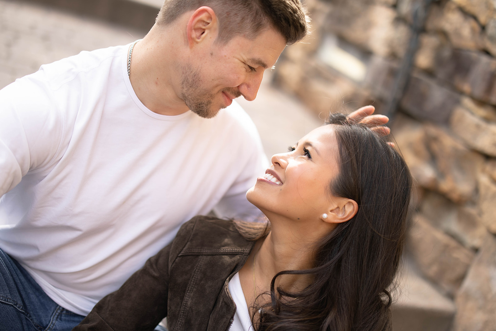 A close-up of a couple sitting together, gazing into each other’s eyes with soft stone textures behind them.