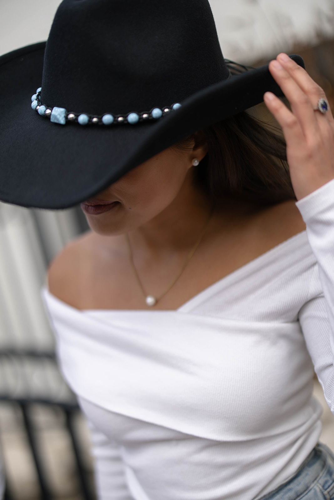 A woman adjusts her black hat, showing a pearl necklace and off-the-shoulder top in a stylish close-up.