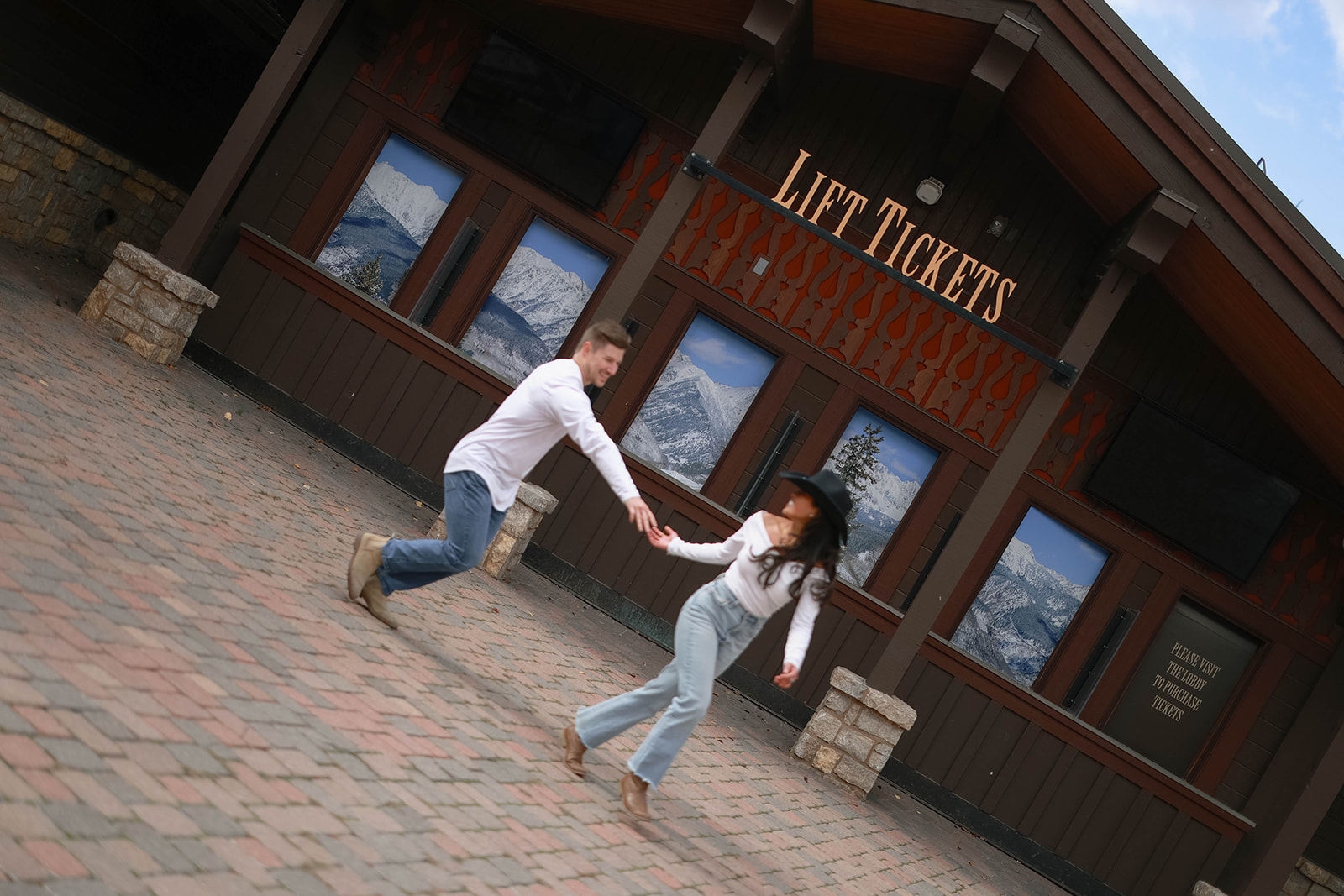 A playful, motion-blurred moment of a couple holding hands and running in front of a wooden lift-ticket building with snowy mountain photos in the windows.