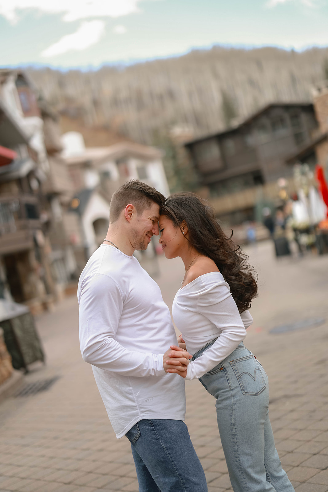 A couple leaning forehead-to-forehead in the center of Vail Village in Colorado, surrounded by alpine-style buildings and early winter mountain views.