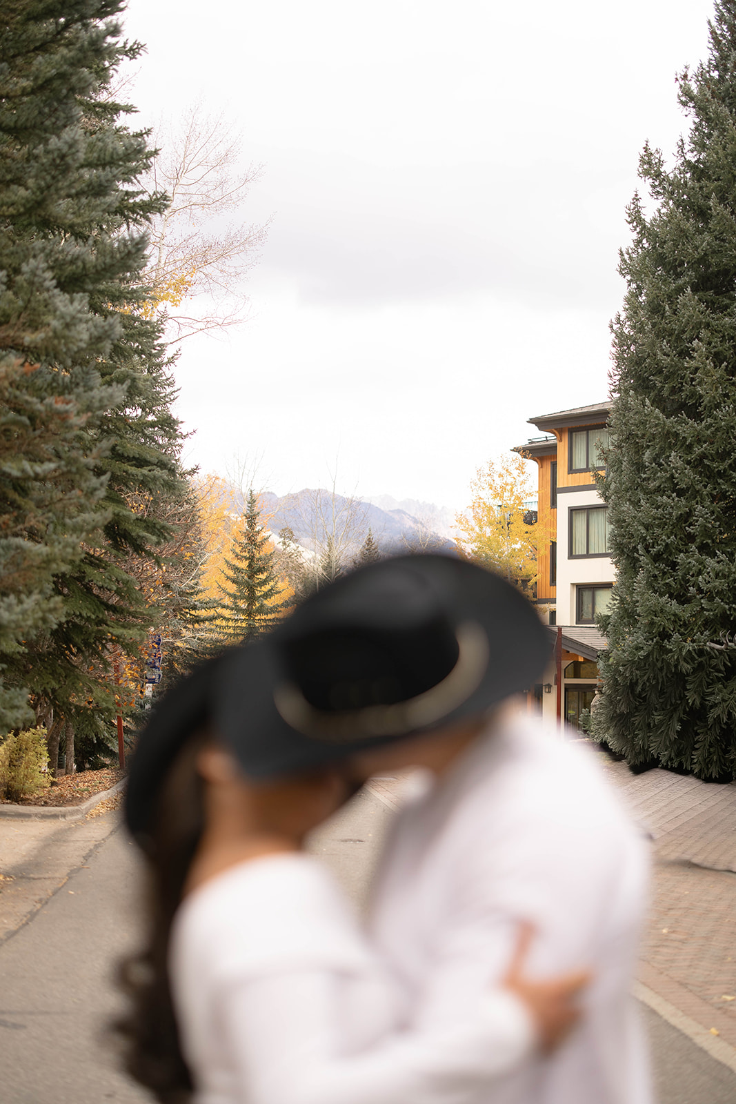 A blurred close-up of a couple kissing while wearing cowboy hats, with fall trees and mountains visible in the background.