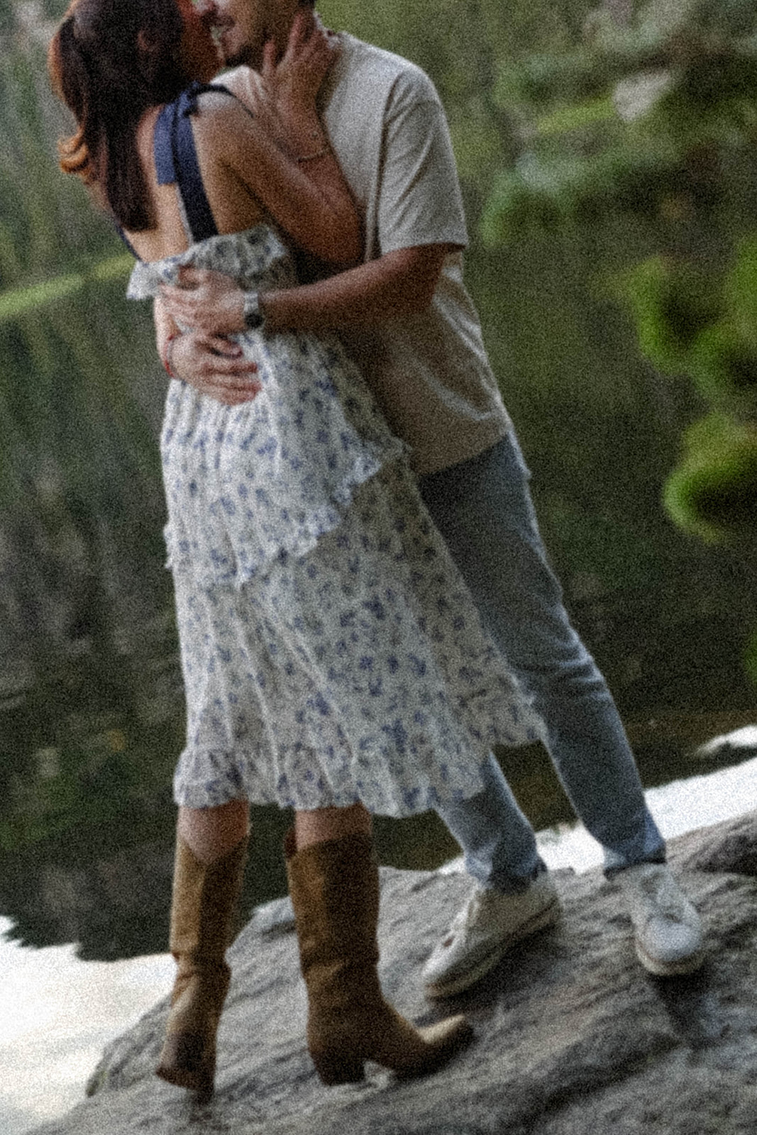A romantic couples photoshoot kiss by the lake, her floral dress flowing as they stand together on the rocks.