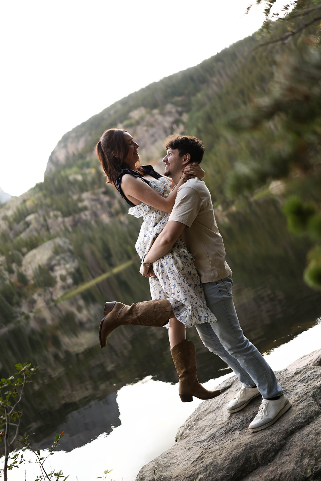 A man lifts his partner beside a mountain lake, both smiling as the sun reflects off the water.