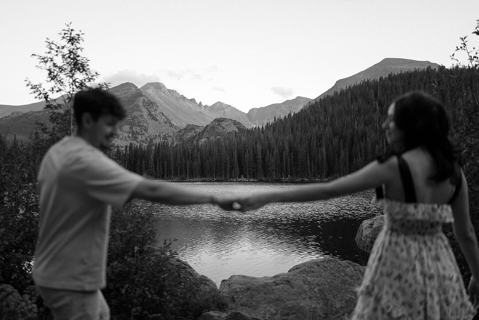 A black-and-white photo captures a couple reaching for each other across a mountain lake, framed by pine trees and distant peaks.