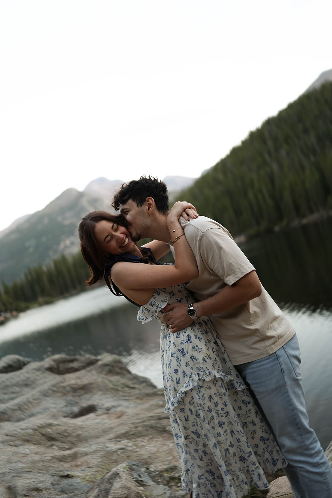 Laughter fills this lakeside couples photoshoot as he wraps his arms around her, both smiling with the mountains softly fading in the distance.