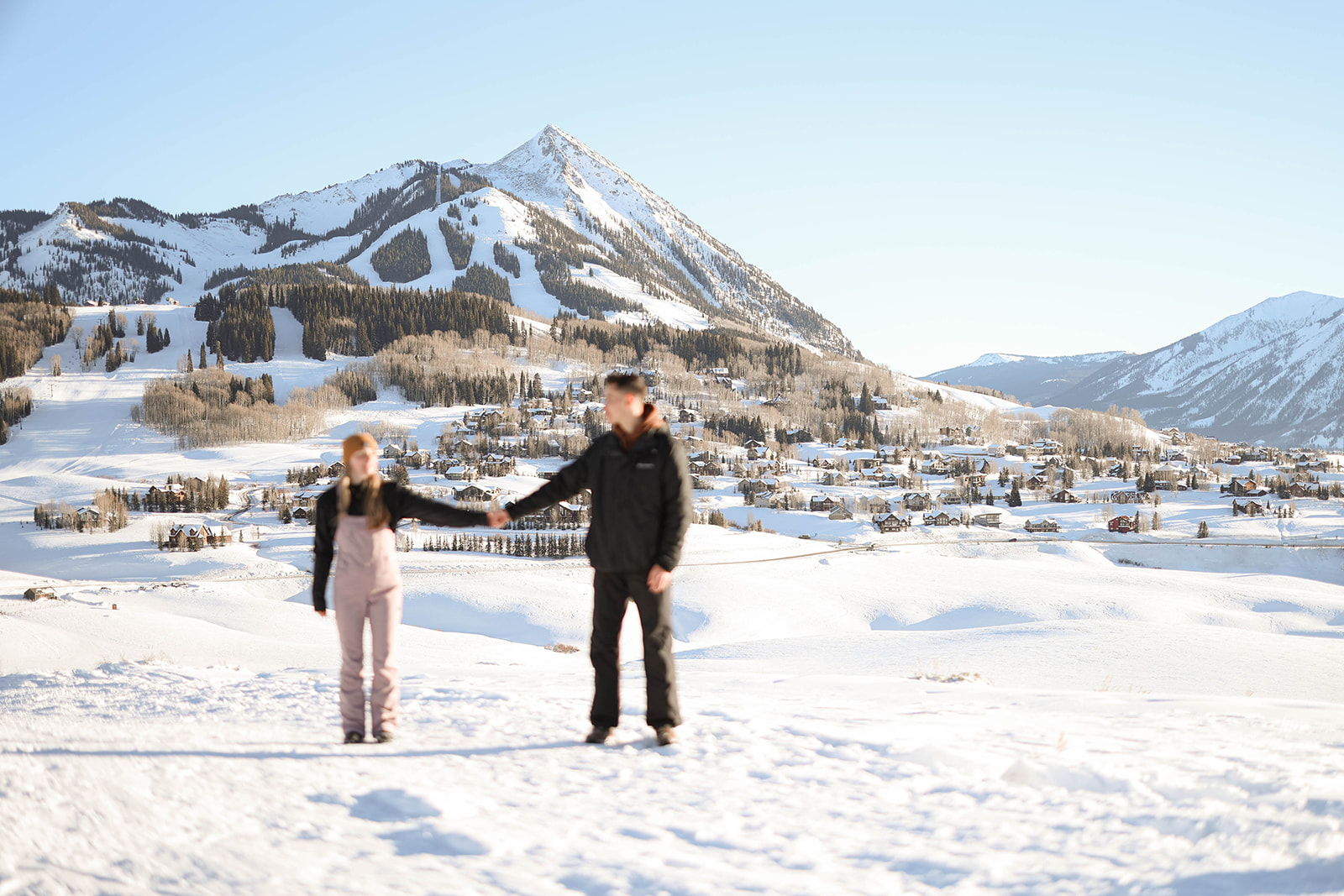 A playful couples photoshoot moment in the Colorado mountains, where two people hold hands across the snow with ski slopes in the background.