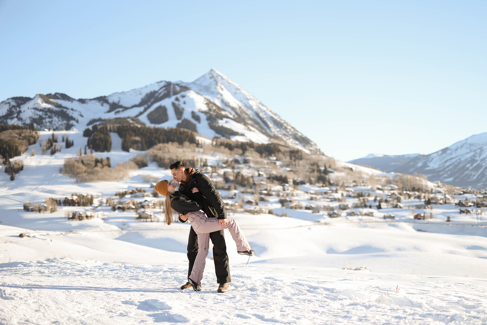 A man dips his partner as they laugh, snow-covered mountains rising in the distance under clear blue skies.