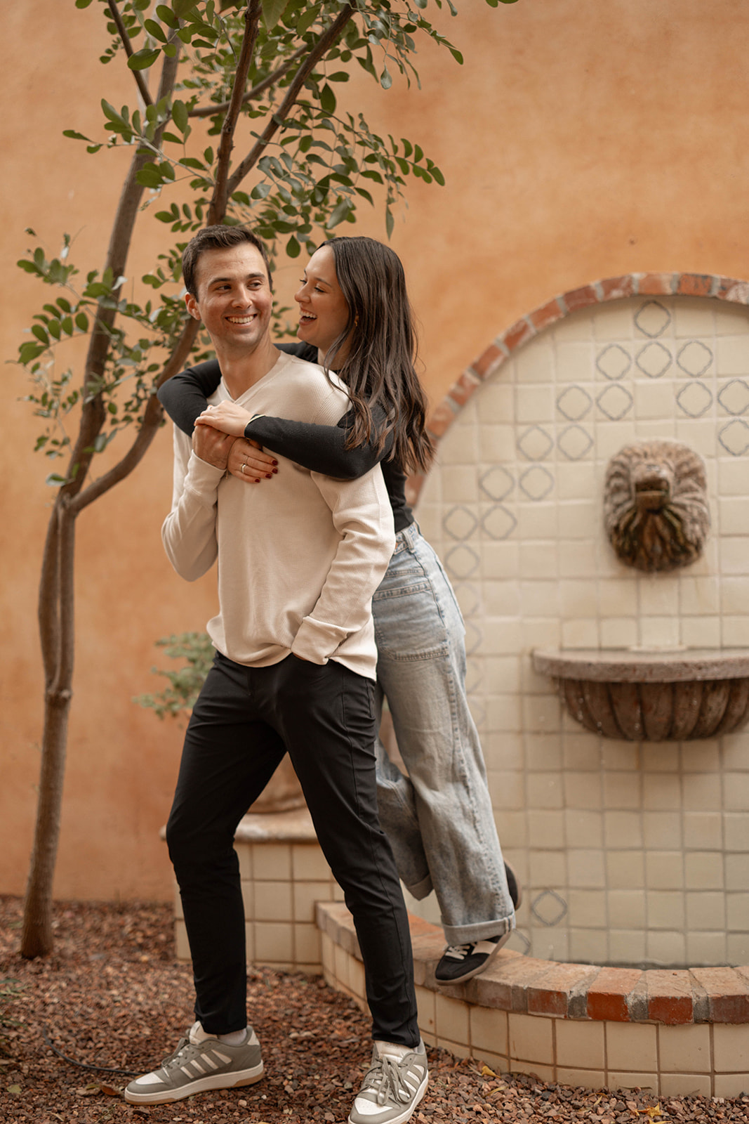 A playful couples photoshoot moment where she jumps on his back, laughing together against a warm terracotta wall.