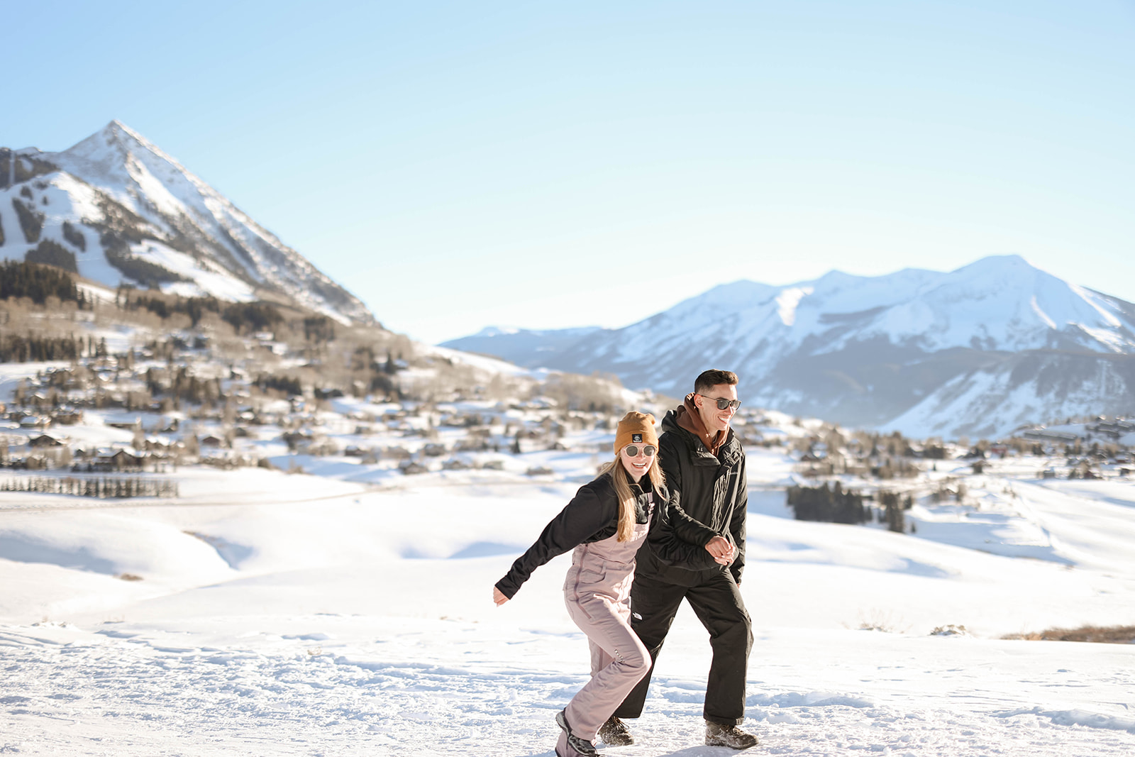 Holding hands and smiling, the couple walks through a snowy mountain town, sunshine glinting off the slopes around them.