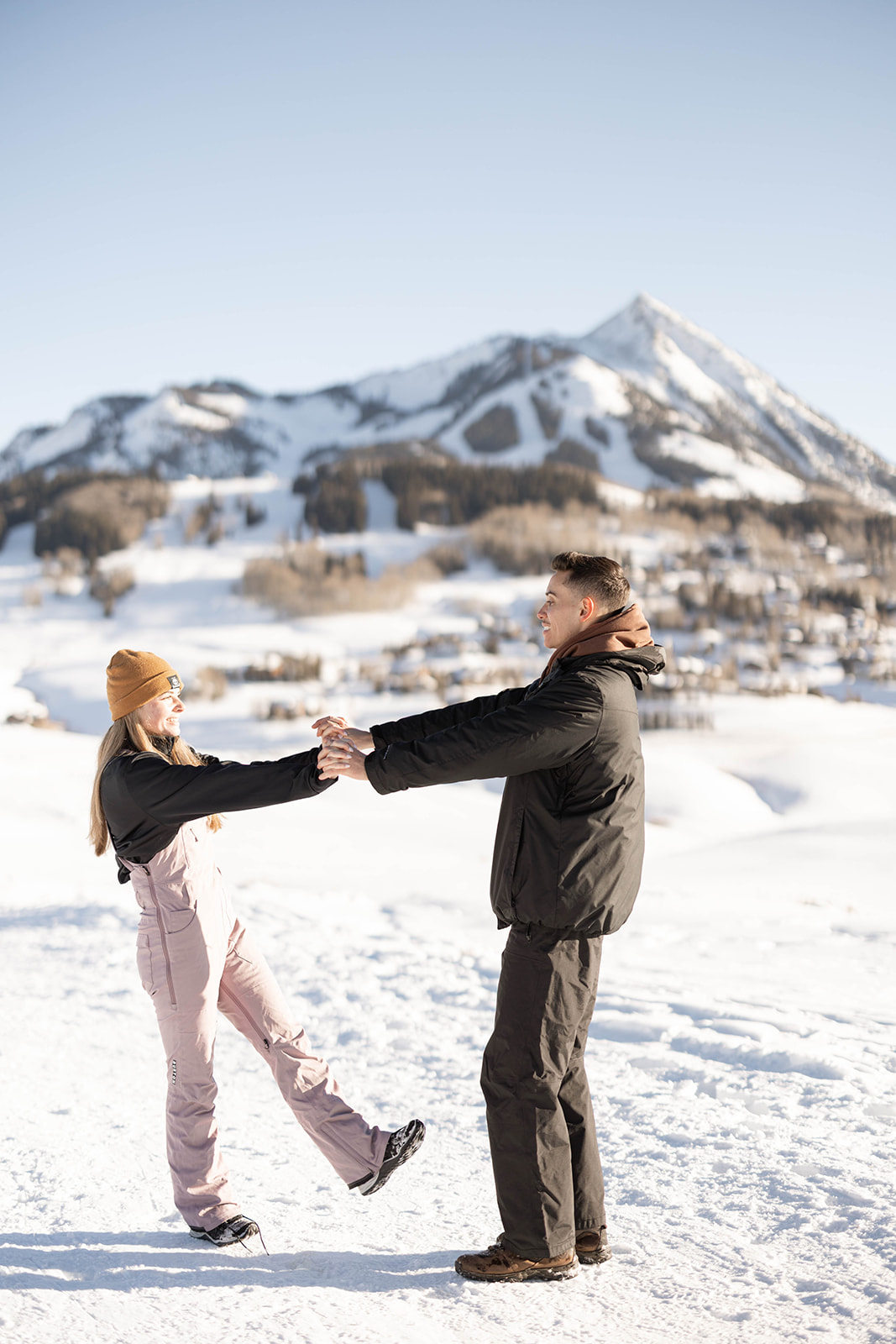 The couple holds hands playfully in the snow, smiling beneath a clear blue sky and mountain peaks.