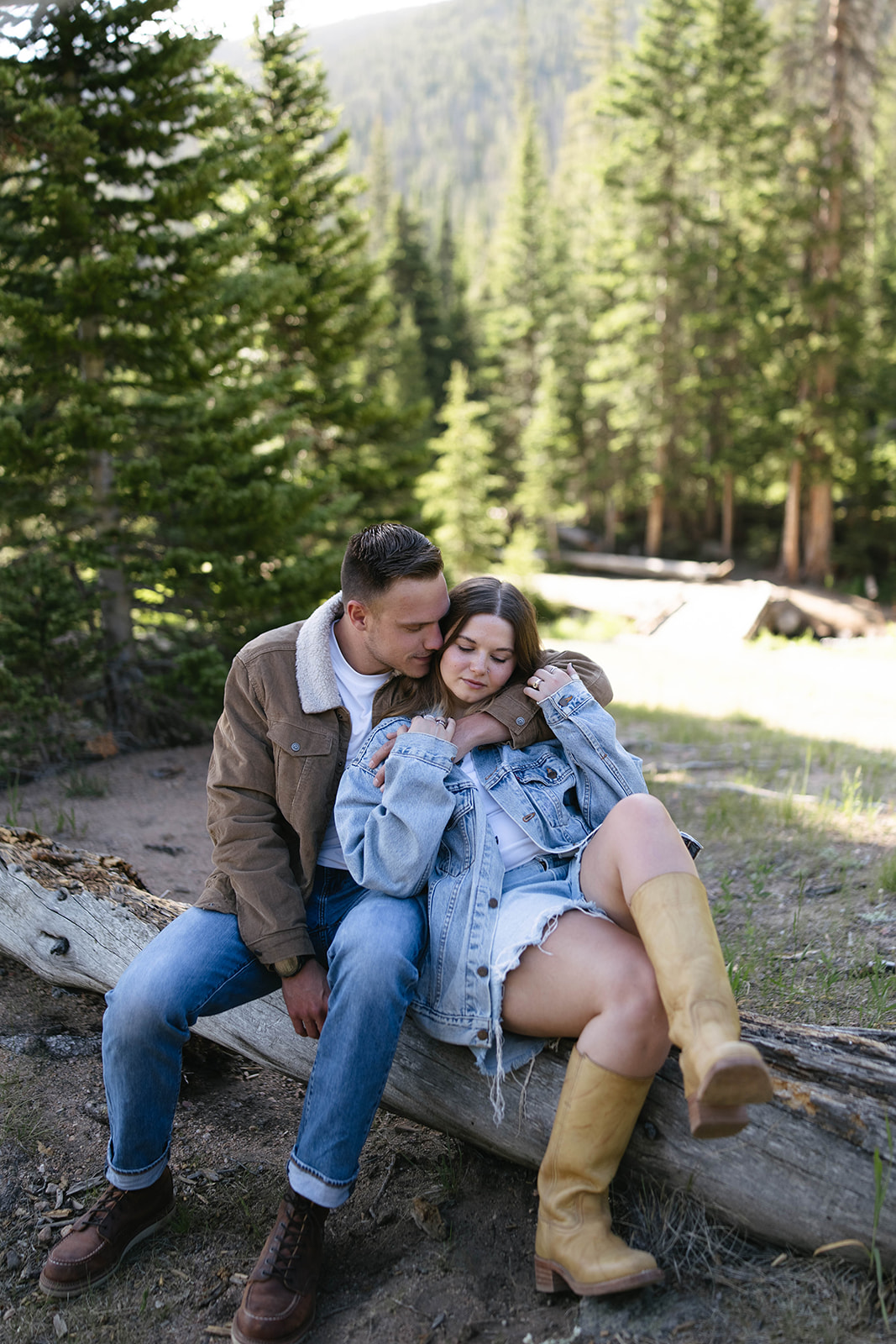 The couple snuggles on a fallen tree surrounded by tall evergreens, their denim and boots adding a cozy, rustic couples photoshoot vibe.
