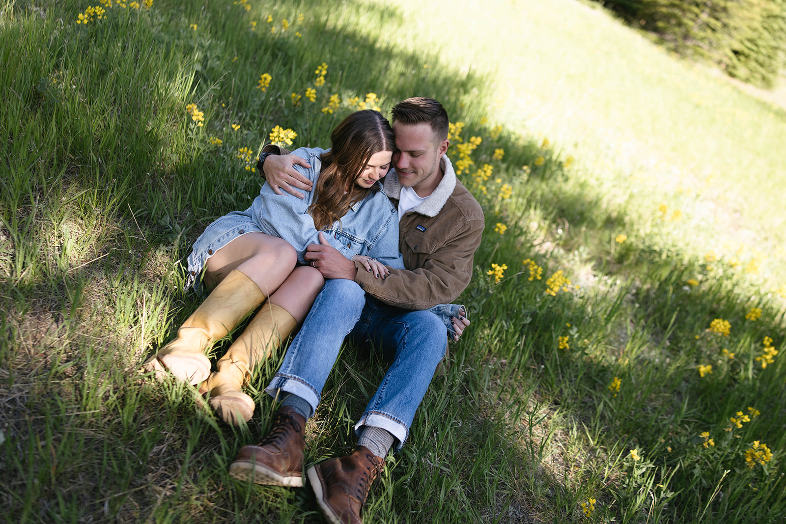 The couple cuddles in a field of yellow wildflowers, wrapped in denim and soft sunlight, a laid-back couples photoshoot that feels effortlessly intimate.