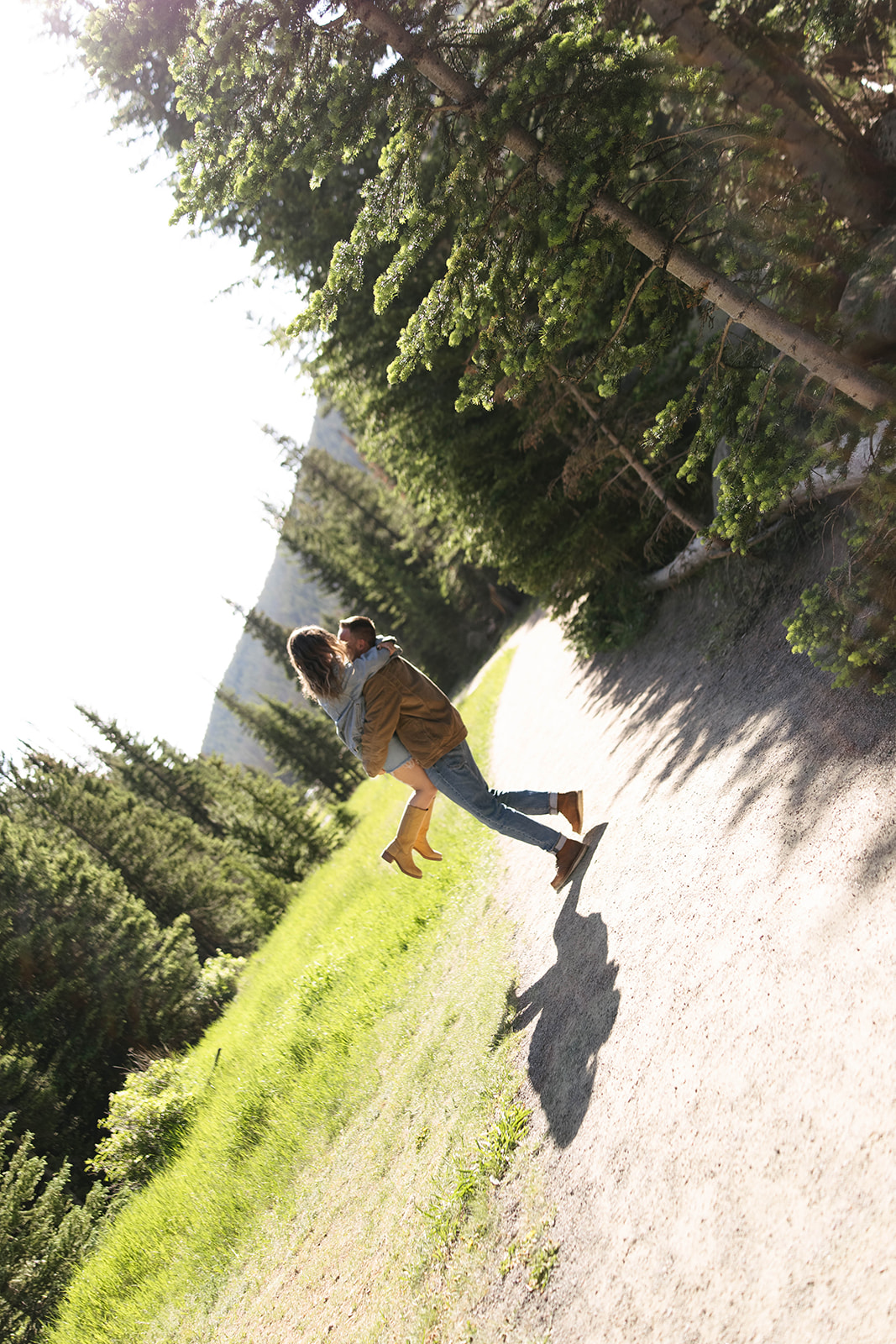 The man lifts his partner into the air on a sunlit forest trail, both laughing as the light filters through tall trees.