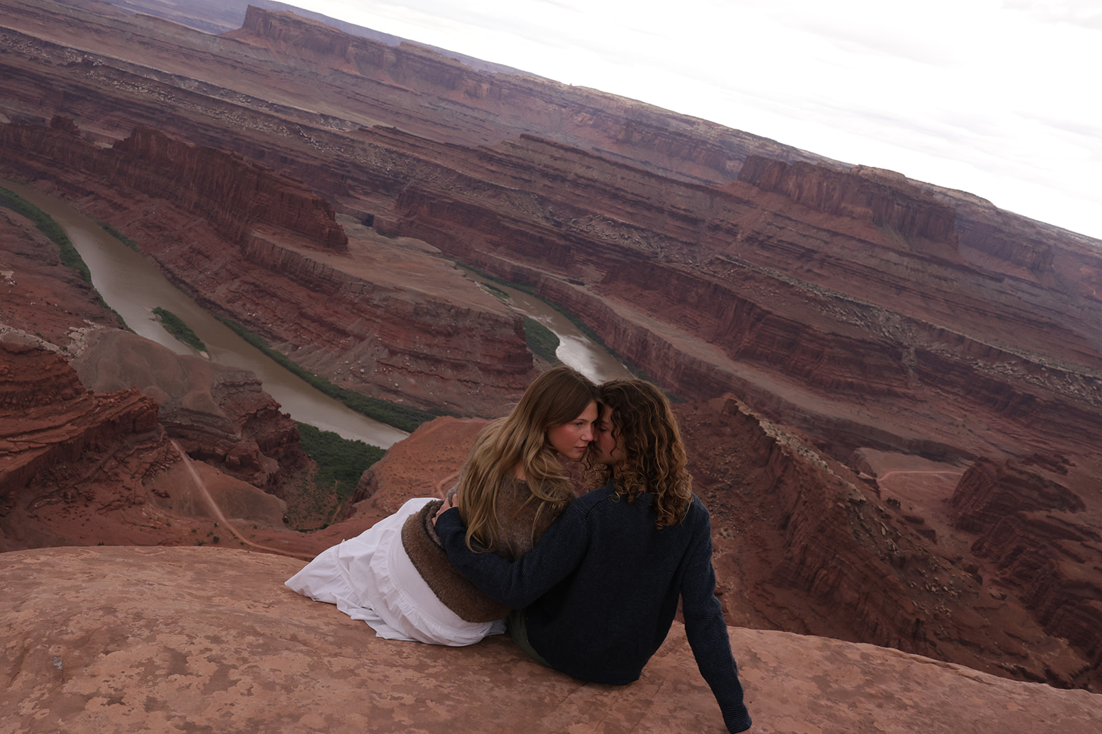 Two people share a quiet couples photoshoot moment at the edge of a canyon, wrapped in sweaters as they look out over winding red rock cliffs.