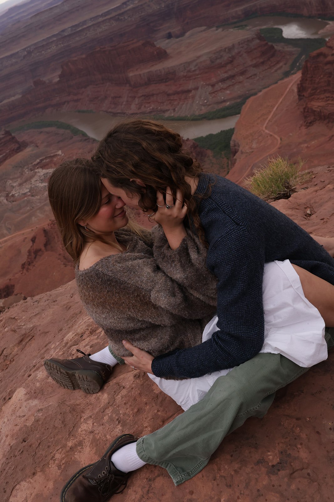 A cozy, intimate scene of a couple sitting close together on the edge of a red rock canyon, sharing a quiet moment above the winding river below.