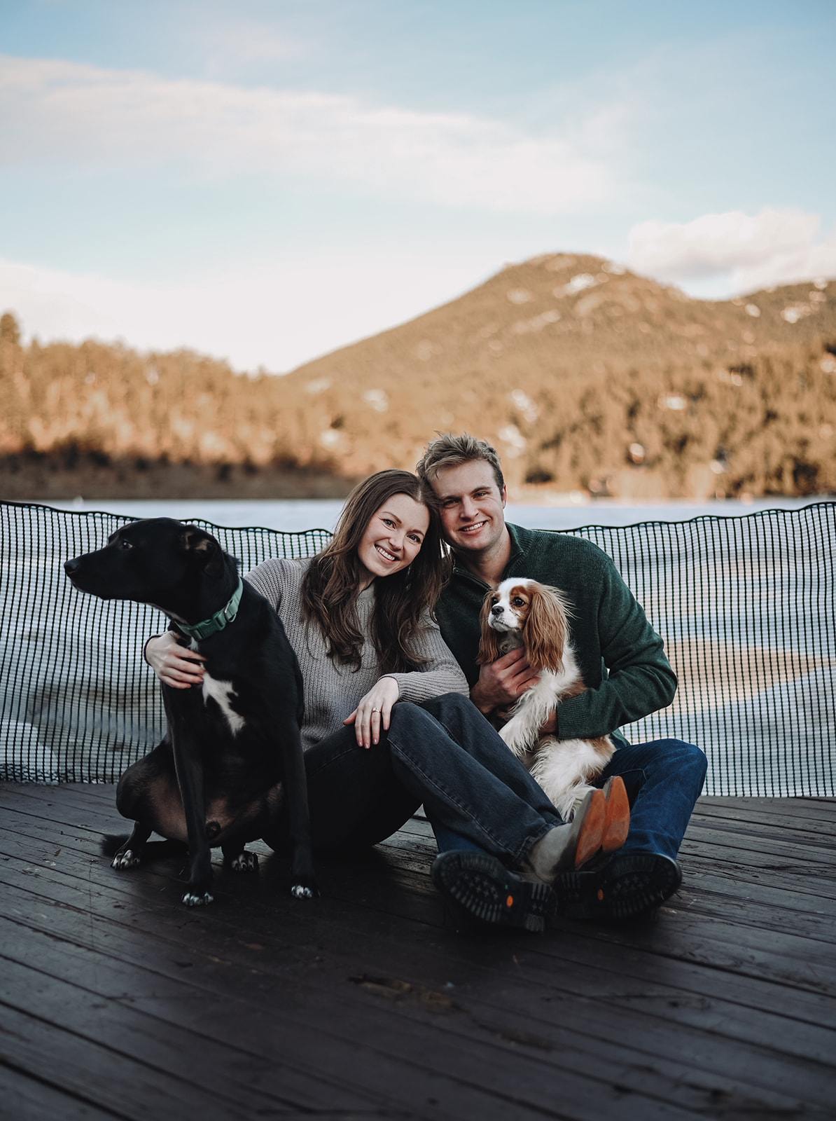 A cozy couples photoshoot moment with a pair sitting on a dock beside their two dogs, surrounded by mountain views and golden light.