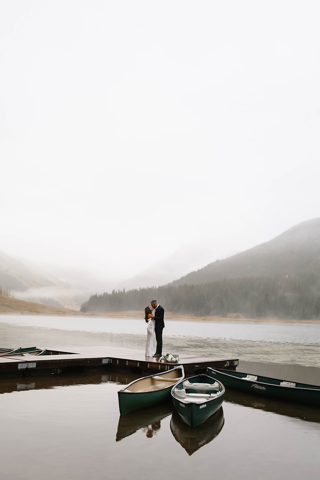 The newlyweds share a quiet kiss on the dock, surrounded by calm lake water and golden autumn colors in the distance.