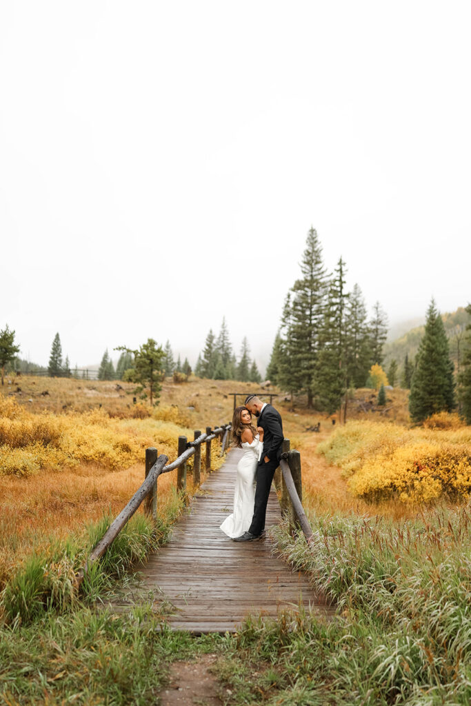 A romantic moment on a rustic wooden bridge framed by golden fall foliage and tall pines in the Colorado mountains.