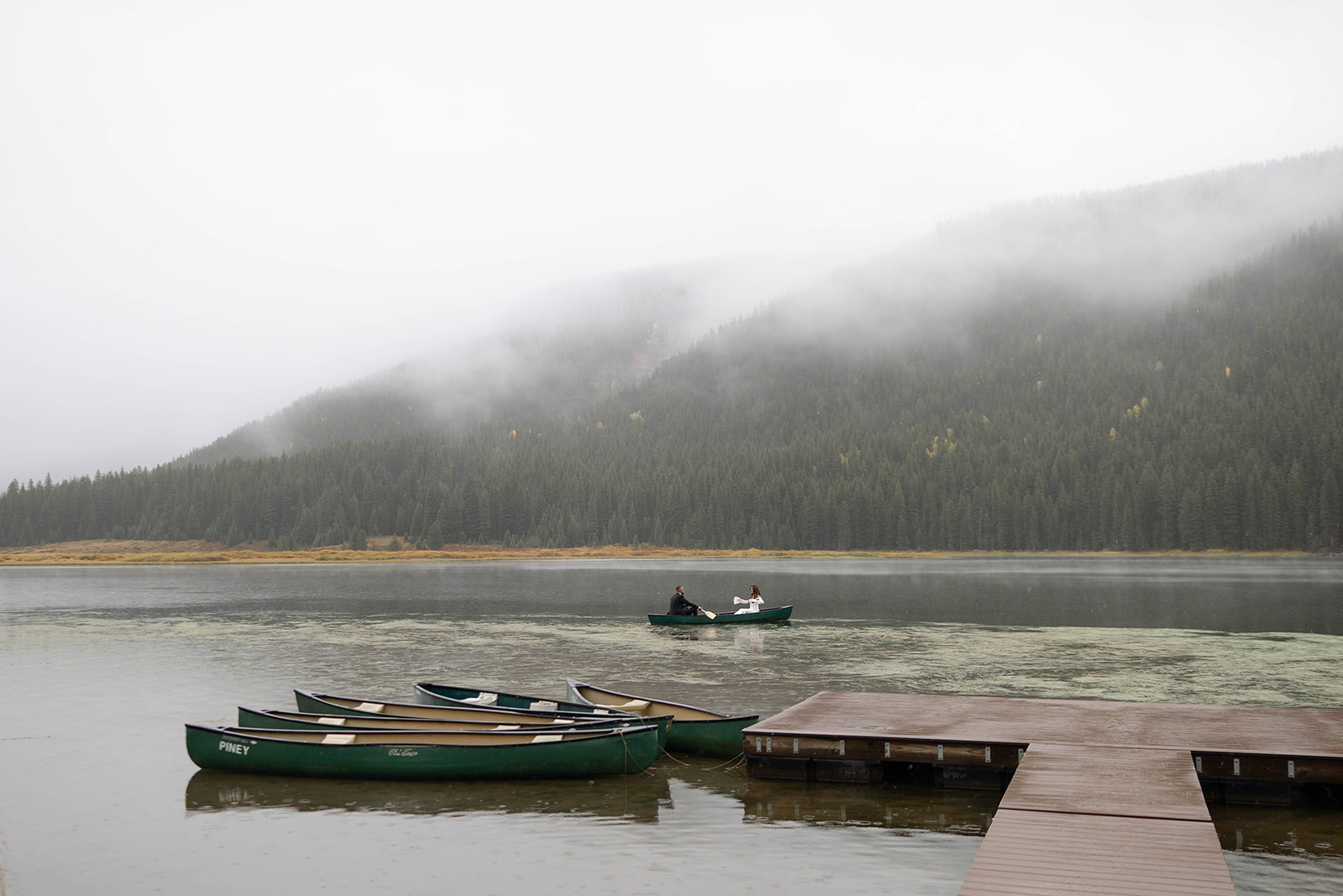 A wide shot of the newlyweds canoeing across the lake, framed by mist, evergreens, and mountain reflections at Piney River Ranch.